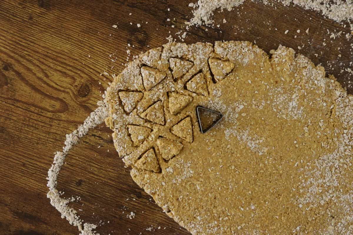 Orange dough with a rough texture rolled out on a floured wooden surface with small triangular shapes cut out of it and a triangular shaped cookie cutter sitting in it.