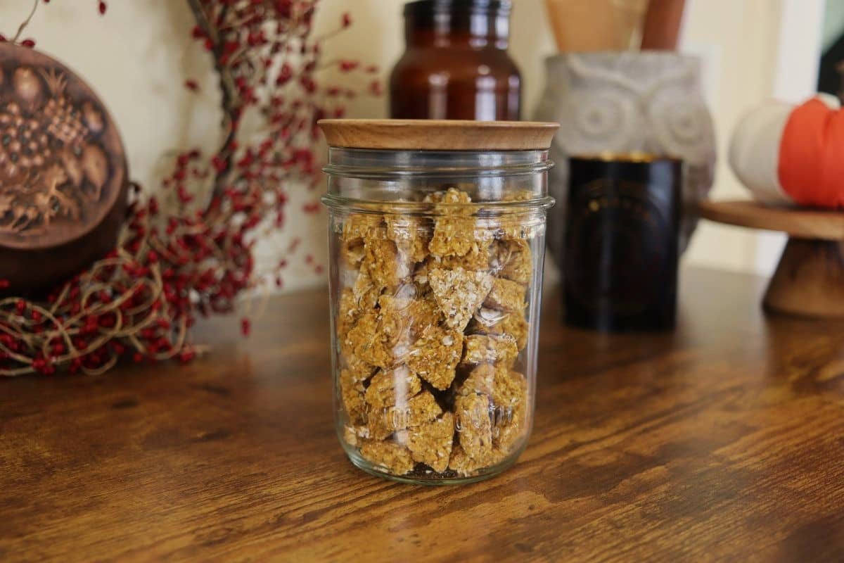 Golden orange, triangular shaped cat treats with a grainy texture in a mason jar with a wooden lid on top sits on a wooden counter with home decor in the background.