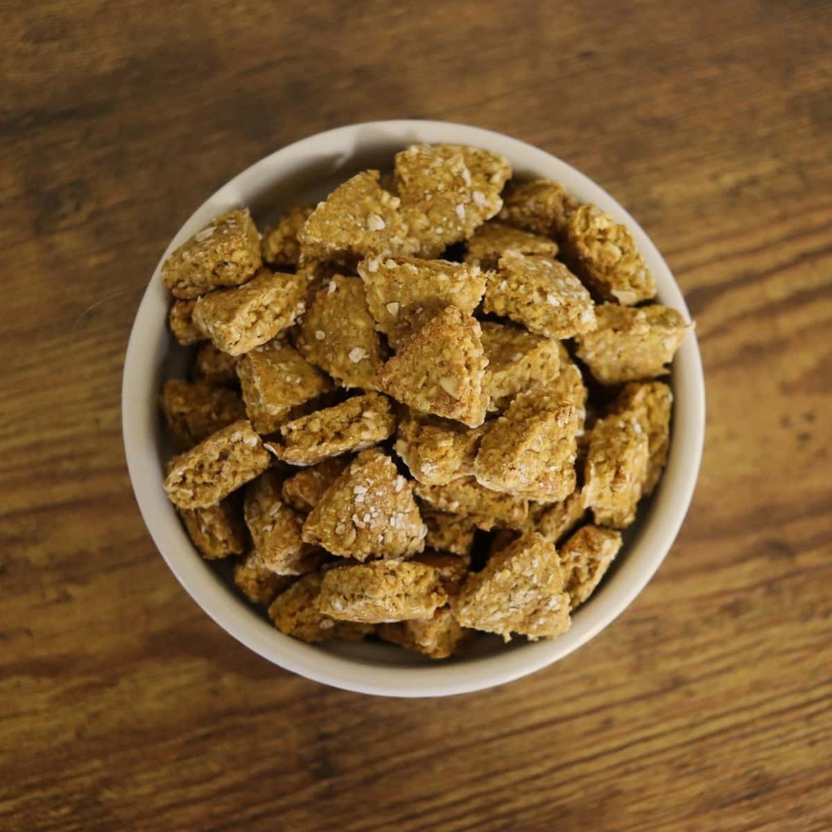 Triangular cat treats with a grainy texture in a white bowl sitting on a wooden surface.