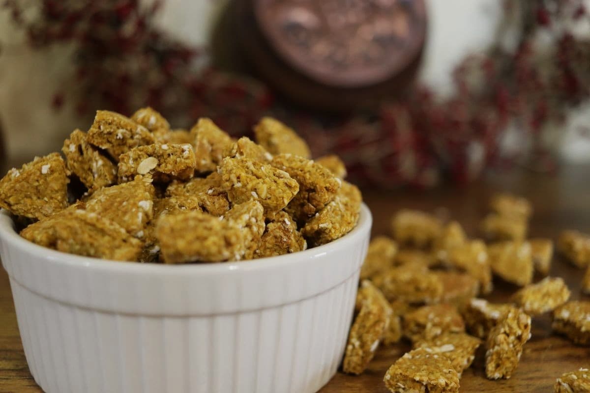 A white bowl filled with golden orange pumpkin cat treats sits on a wooden surface, with more treats scattered beside it. Blurred red berries and a decorative item are in the background.
