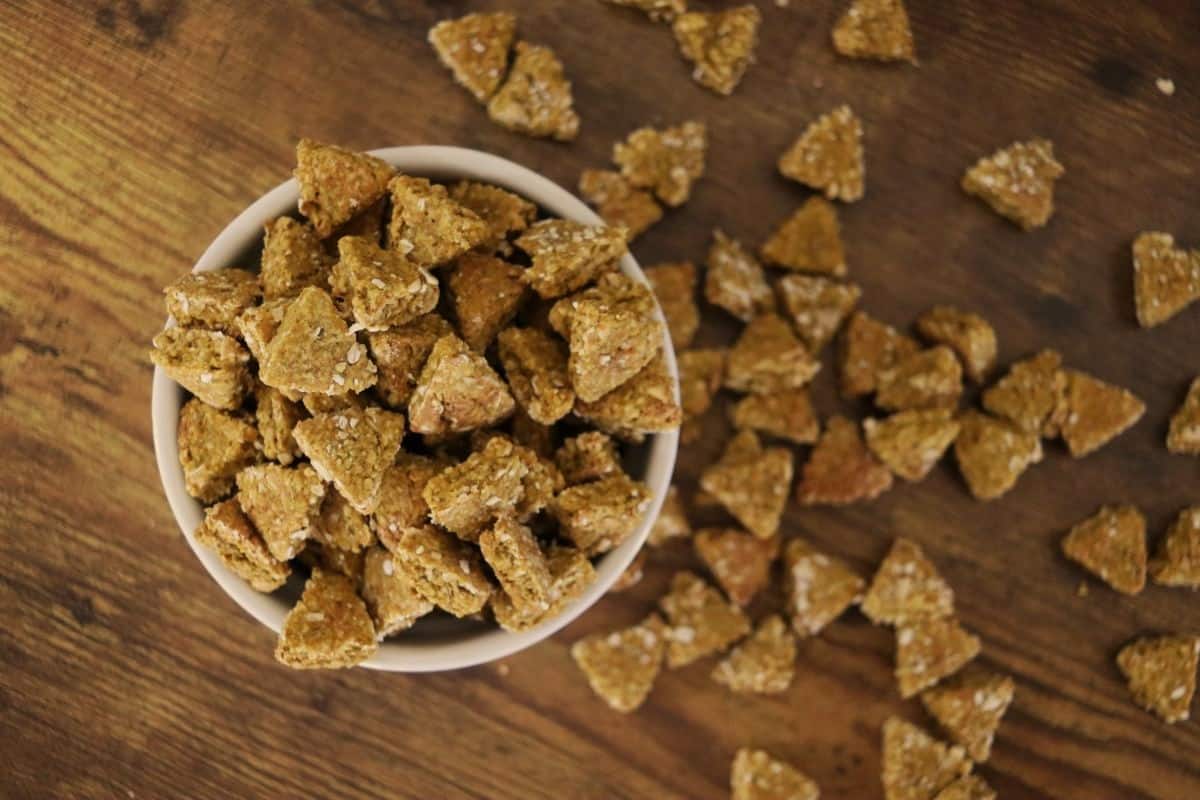 A white bowl filled with golden orange pumpkin cat treats sits on a wooden surface, with more treats scattered beside it. Blurred red berries and a decorative item are in the background.