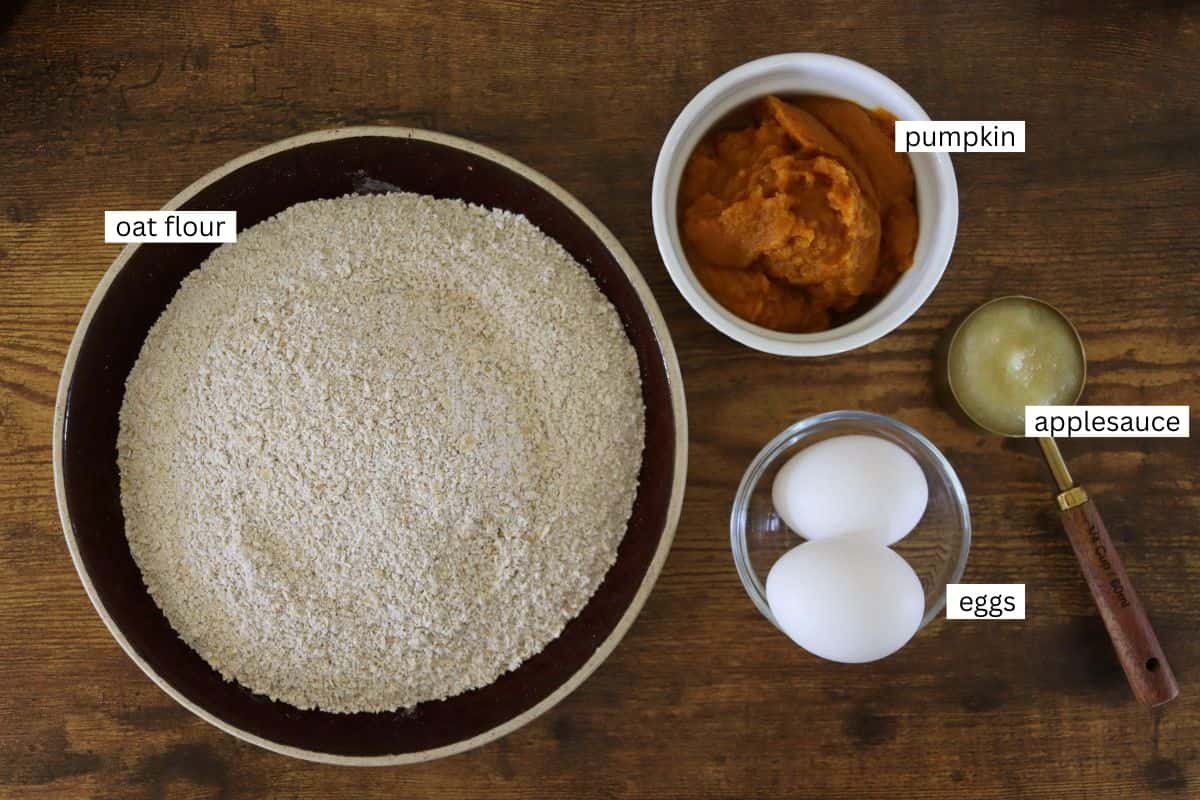 A bowl of oat flour, a small bowl of pumpkin puree, a spoonful of applesauce, and two eggs are arranged on a wooden surface, each labeled with their ingredient name.