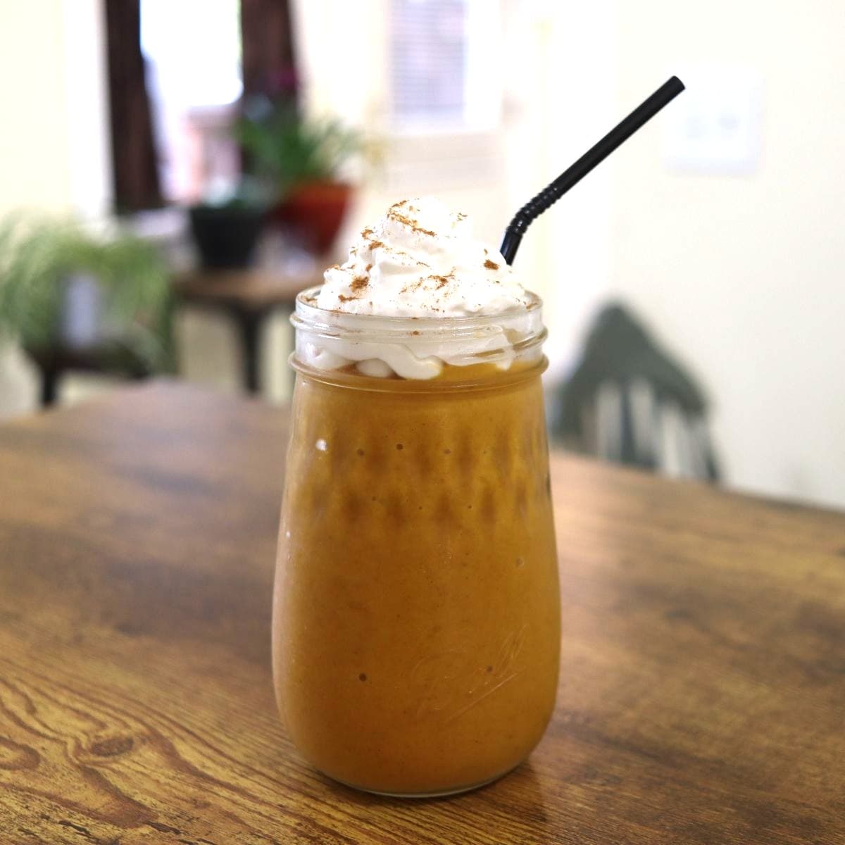 Pumpkin smoothie in a ball flute mason jar topped with whipped cream and a dusting of cinnamon with a black straw in it.