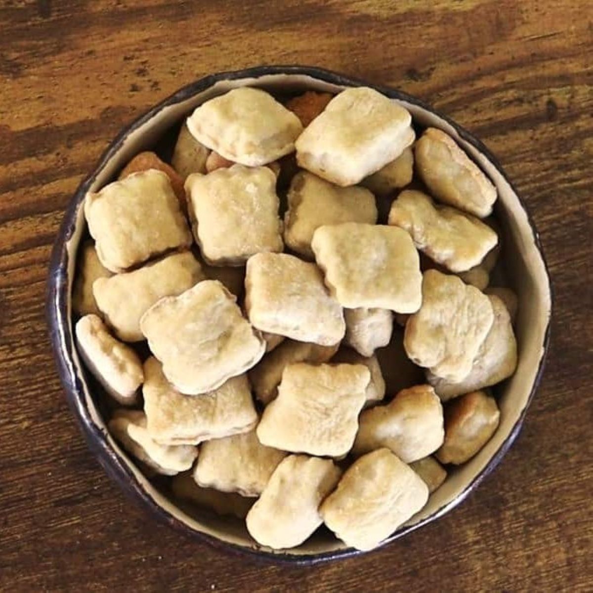 Square crackers in a brown bowl on a wooden table.