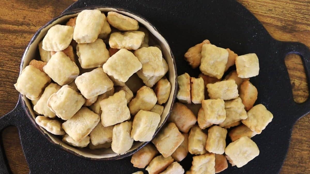 A bowl and a black tray filled with small, square, golden-brown baked crackers, with some spilling out of the bowl onto the tray.