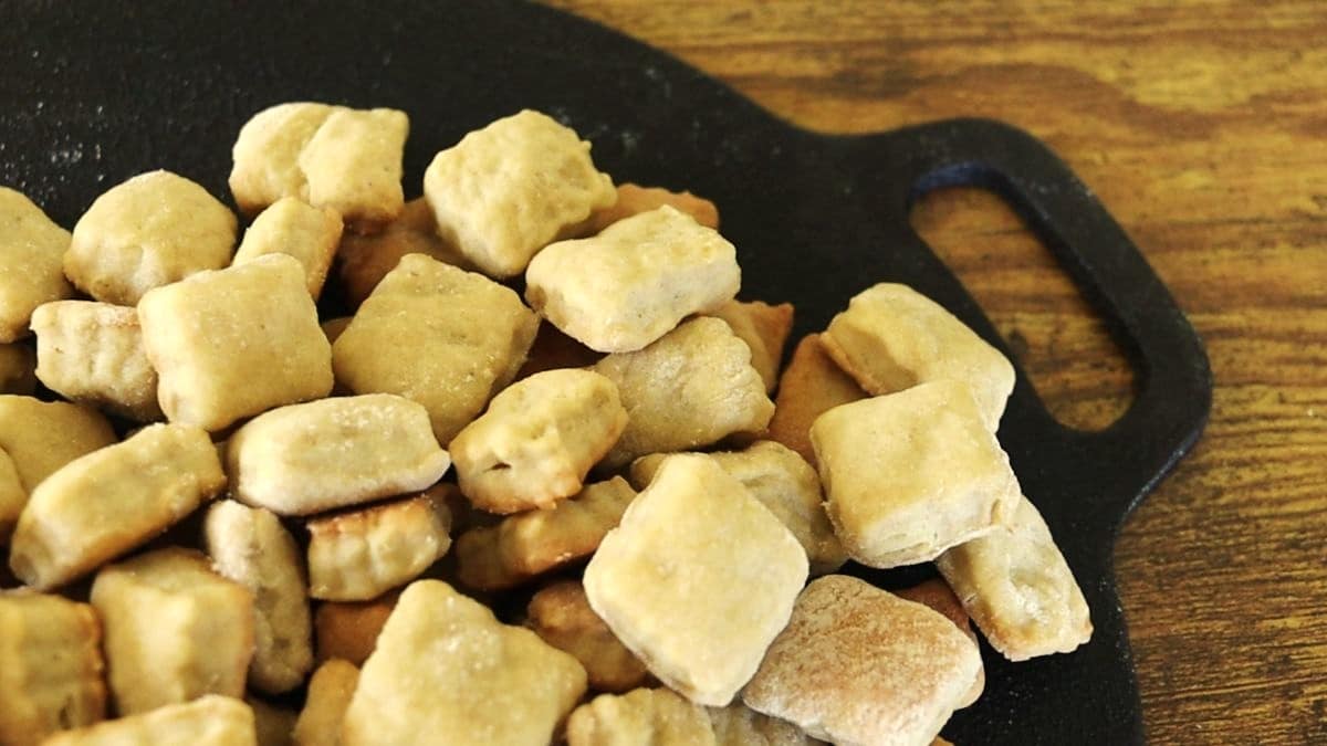 A pile of golden-brown, bite-sized crackers is arranged on a black serving tray with a handle, resting on a wooden surface.