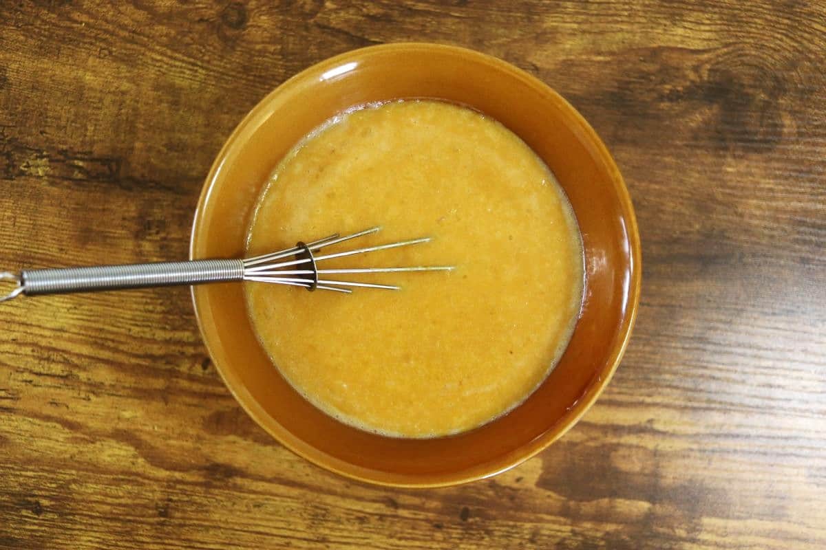 Orange colored batter in a brown bowl with a whisk in it sitting on a wooden surface.