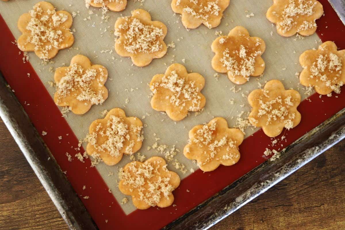 Orange, flower shaped crackers topped with brown sugar sitting on a silicone lined baking sheet.