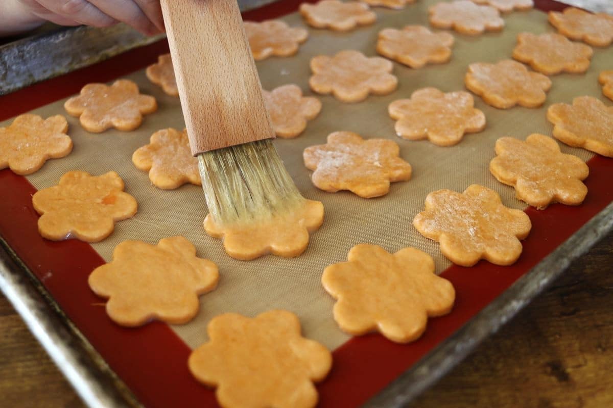 Orange, flower shaped crackers on a silicone lined baking sheet being basted with an egg mixture.