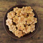 Light orange, flower shaped crackers with sugar topping piled high on a brown plate sitting on a wooden surface.