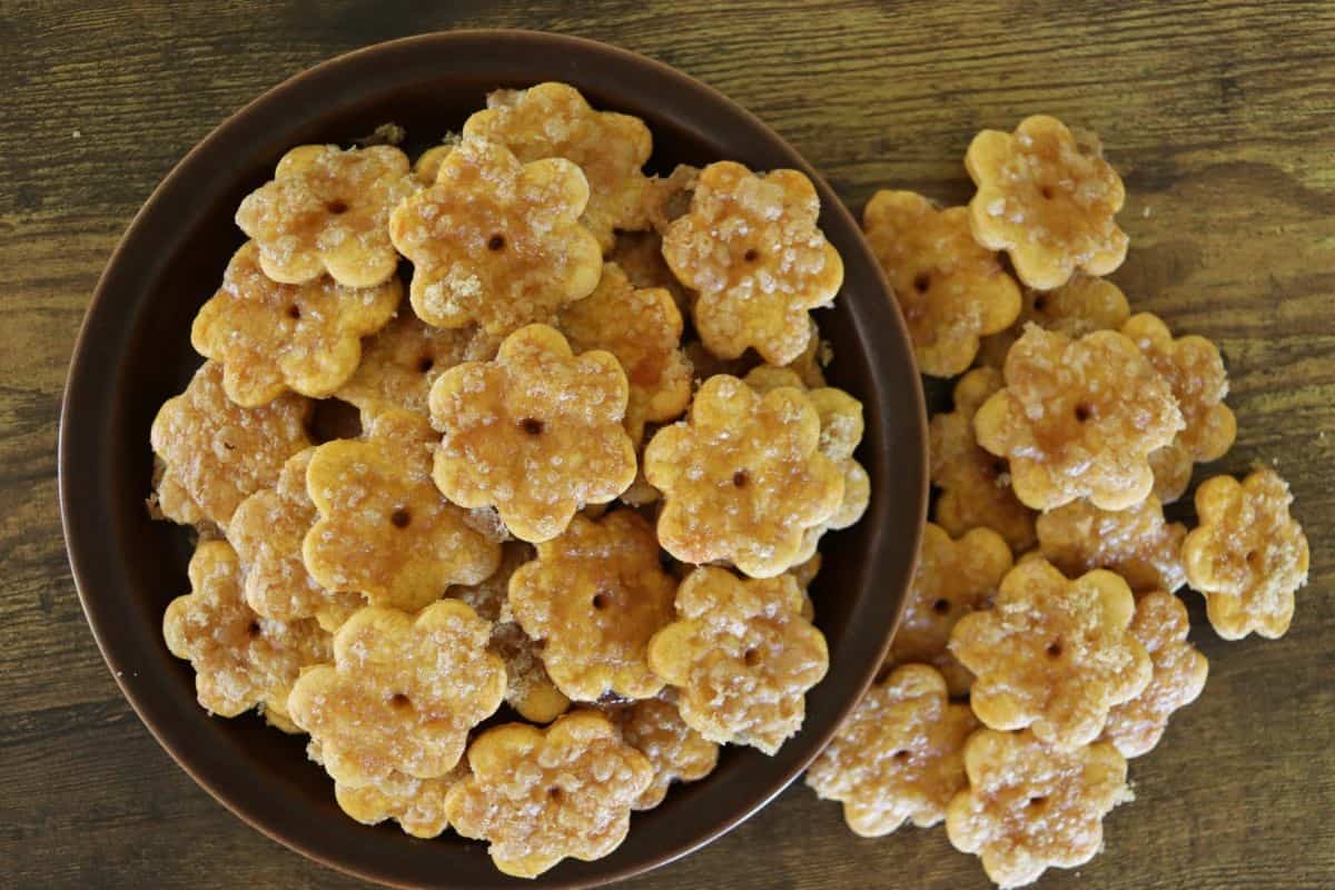 A brown plate filled with flower-shaped, golden-brown crackers sprinkled with sugar, with some crackers scattered on a wooden surface next to the plate.