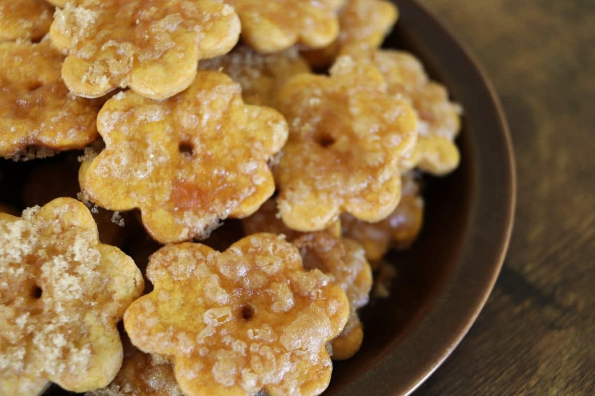 A close-up of flower-shaped crackers with a sugary coating, arranged on a dark plate. The crackers are golden brown and appear freshly baked.