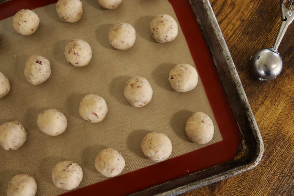 Balls of tan colored dough with small pieces of nuts visible in them lined up on a silicone lined baking sheet.
