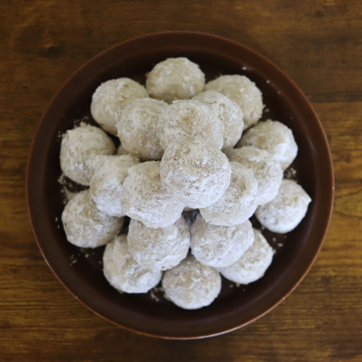 Plate of round cookie balls coated in white powder and piled pyramid style sitting on a wooden surface.