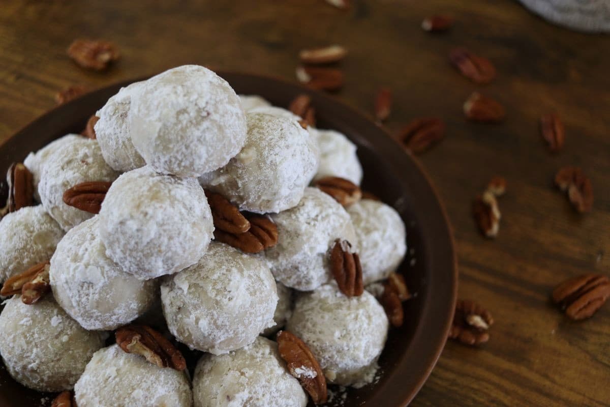 A plate piled with round cookies coated in powdered sugar, garnished with pecan halves, sits on a wooden surface with scattered pecans nearby.