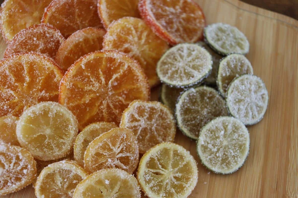 Slices of citrus fruits, including oranges, lemons, and limes, coated in sugar and arranged on a wooden board. The candied slices glisten and display varying shades of orange, yellow, and green.