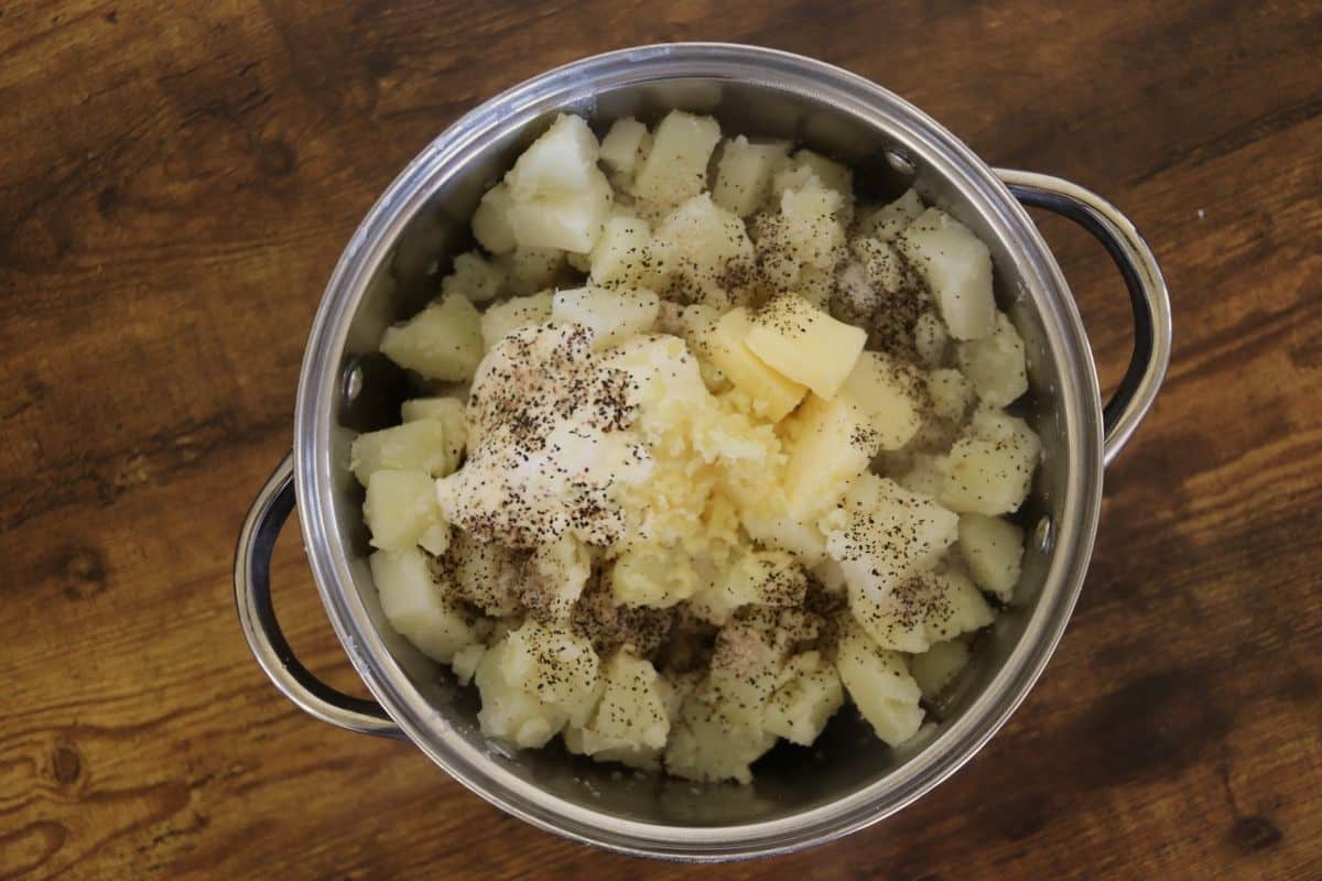 Cooked cut up potatoes in a pot with butter, sour cream, black pepper, and crushed garlic on top of them.