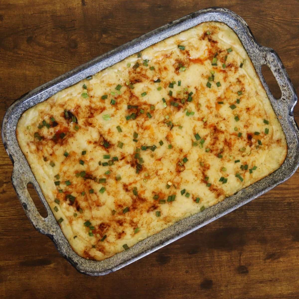 Blue baking dish full of mashed potatoes topped with melted white cheese, paprika, and chopped scallions on top.