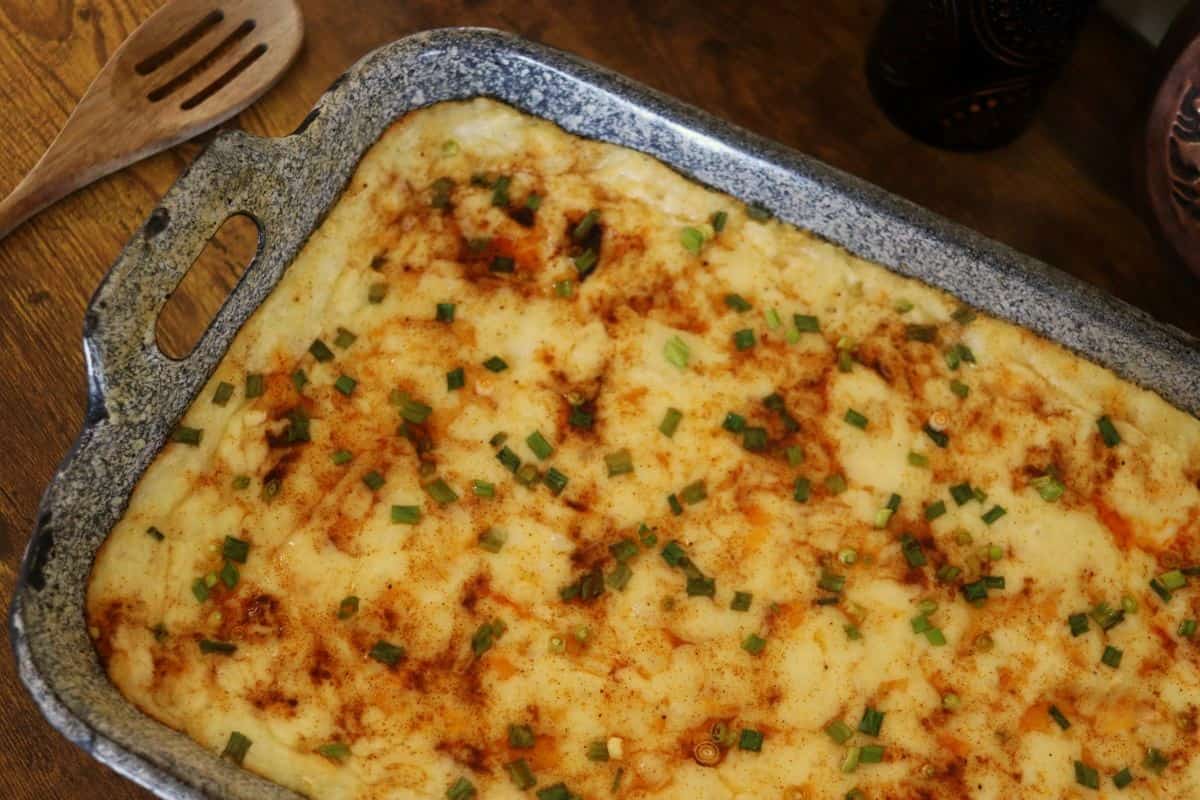 A baking dish filled with mashed potatoes, sprinkled with chopped green onions and paprika, sits on a wooden table next to a wooden slotted spoon.