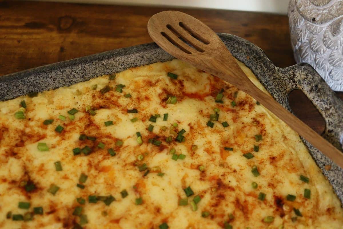 A casserole dish of baked mashed potatoes topped with chopped chives and paprika sits on a wooden table; a wooden slotted spoon rests on the edge of the dish.