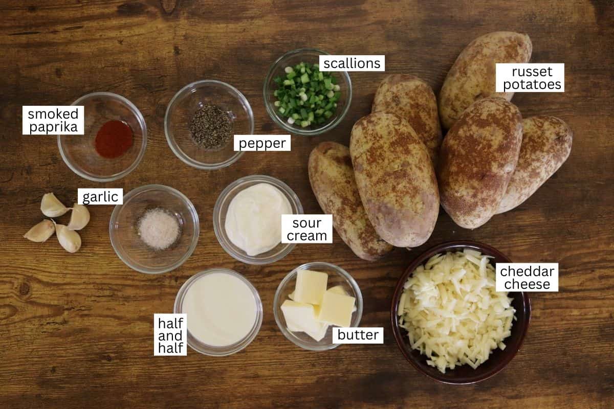 A wooden table with labeled ingredients for mashed potatoes: russet potatoes, scallions, smoked paprika, pepper, garlic, sour cream, cheddar cheese, half and half, and butter, each in separate bowls.
