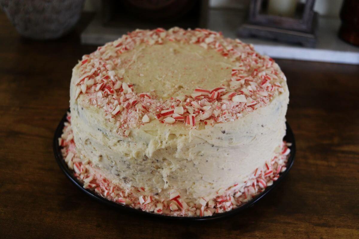 A round cake with white frosting is topped and surrounded by crushed peppermint candy pieces, displayed on a dark plate on a wooden surface.