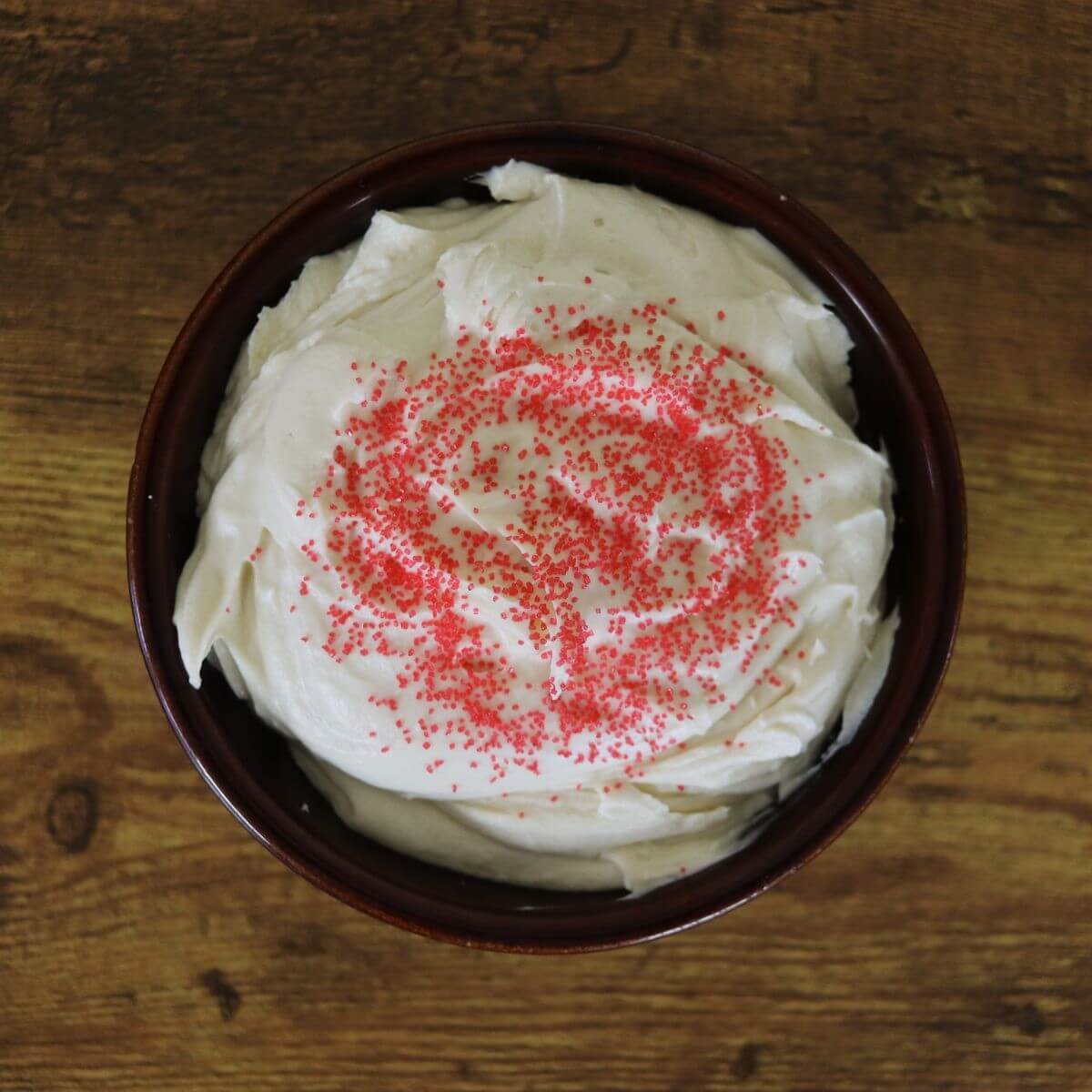 Creamy white dip topped with red sugar crystals in a brown bowl sitting on a wooden surface.