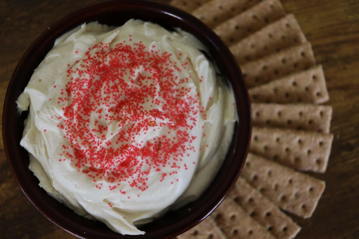 A bowl of creamy white dip topped with red sprinkles, surrounded by rectangular graham crackers arranged in a fan shape on a wooden surface.