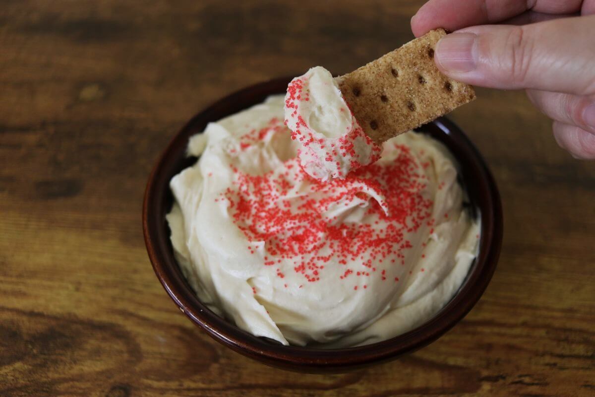 A hand dips a graham cracker into a bowl of creamy white dip topped with red sprinkles, set on a wooden surface.