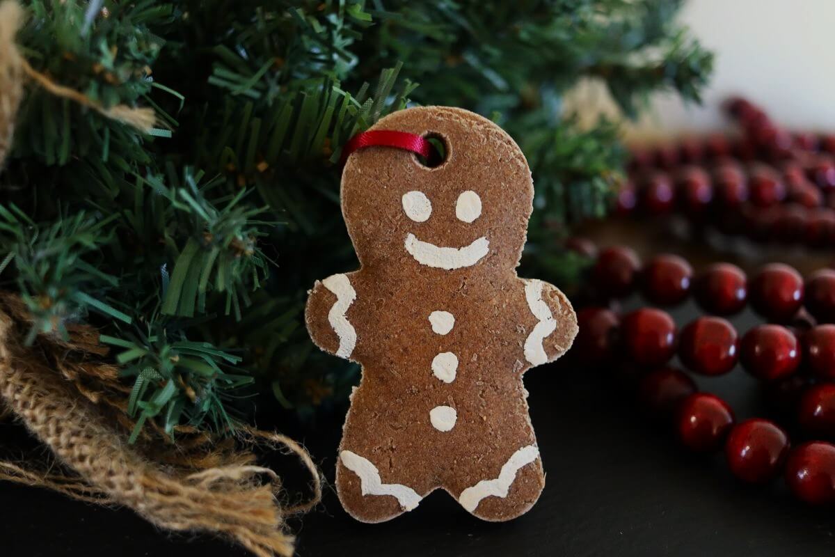 A gingerbread man ornament with white paint details and a red ribbon hangs on a pine branch, with red bead garland and burlap cloth nearby.