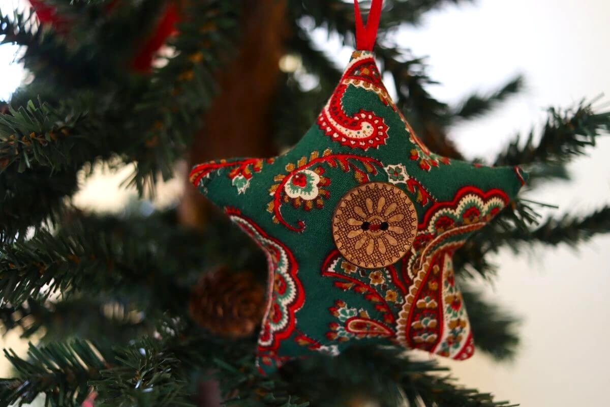 A fabric star-shaped ornament with a red, green, and white paisley pattern and a wooden button hangs on a Christmas tree branch.