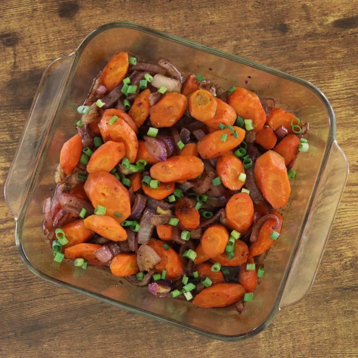 Square baking dish of carrots and red onions topped with chopped scallions sitting on a wooden surface.