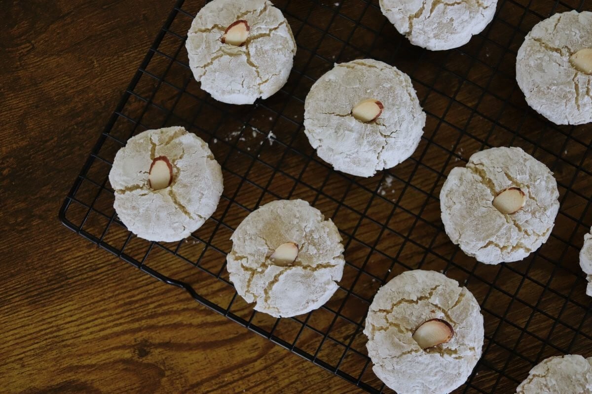 Flat round cookies covered in powdered sugar with an almond slice in the center of them sitting on a wire rack over a wooden table.