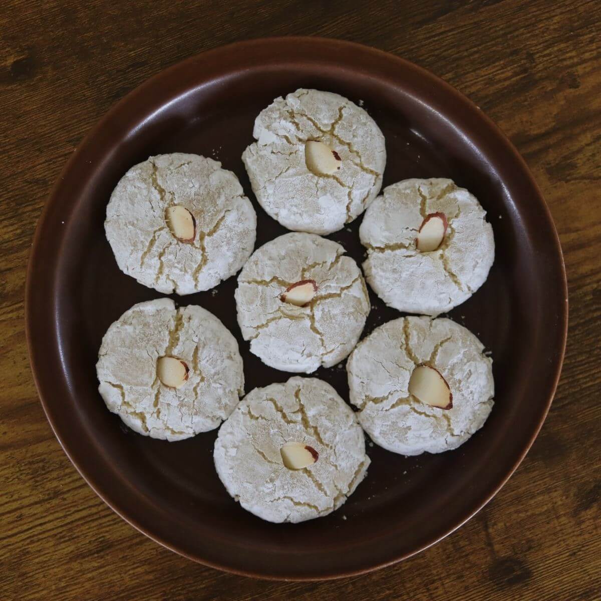 Round cookies coated in powdered sugar with an almond sliver in the center of them arranged on a round plate.