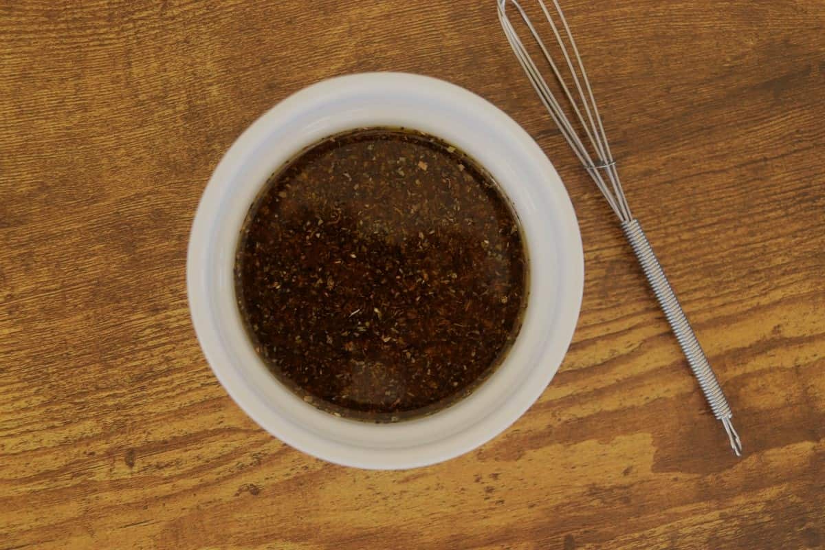 Dark brown sauce with herbs visible in it in a small white dish sitting on a wooden table with a whisk next to it.