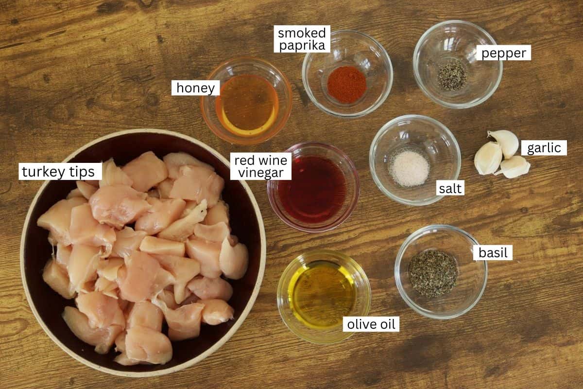 A bowl of raw cubed turkey tips sits on a wooden surface, surrounded by small bowls containing smoked paprika, pepper, garlic, salt, basil, olive oil, red wine vinegar, and honey. Each ingredient is labeled.