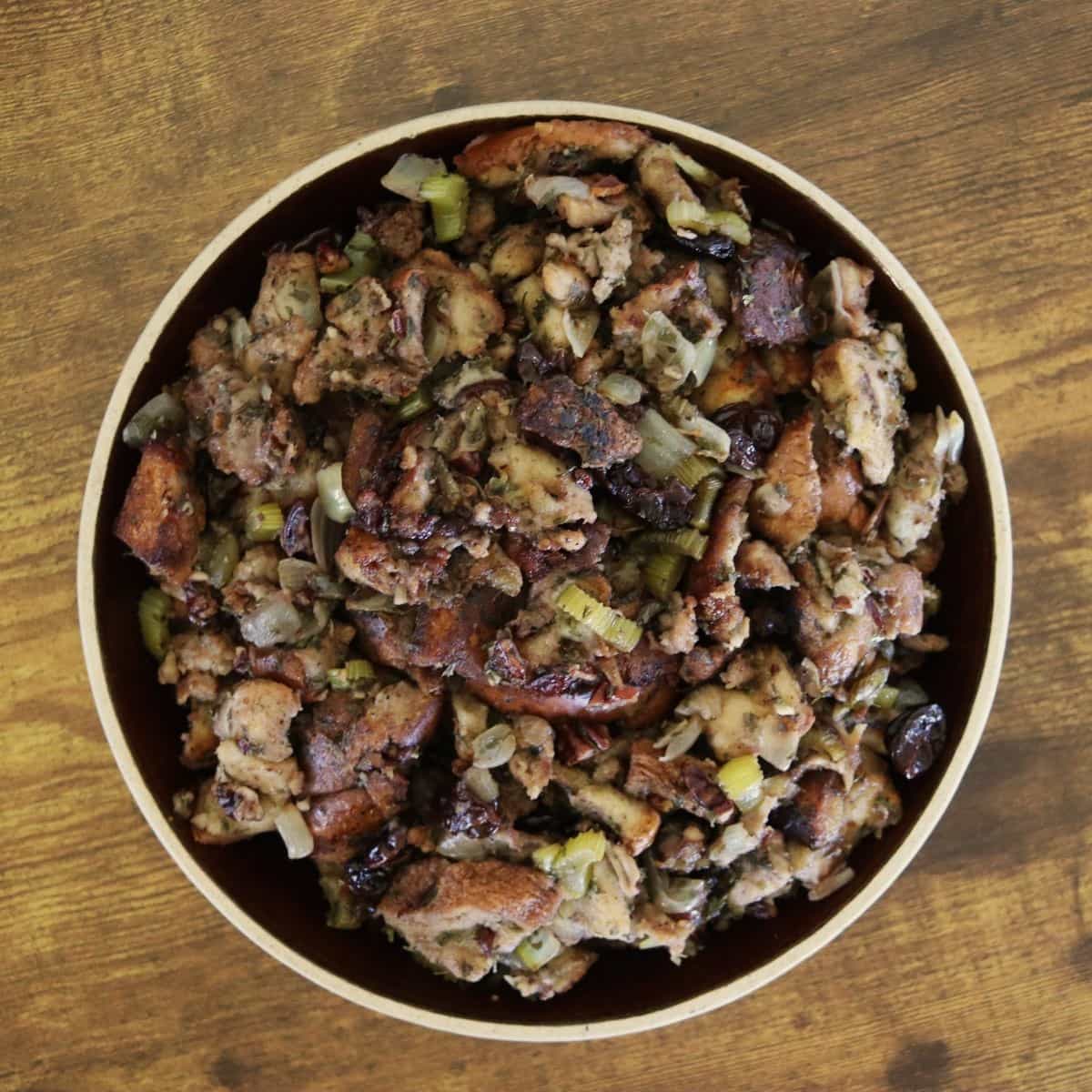 A bowl filled with homemade bread stuffing containing chunks of bread, vegetables, and herbs sits on a wooden surface.