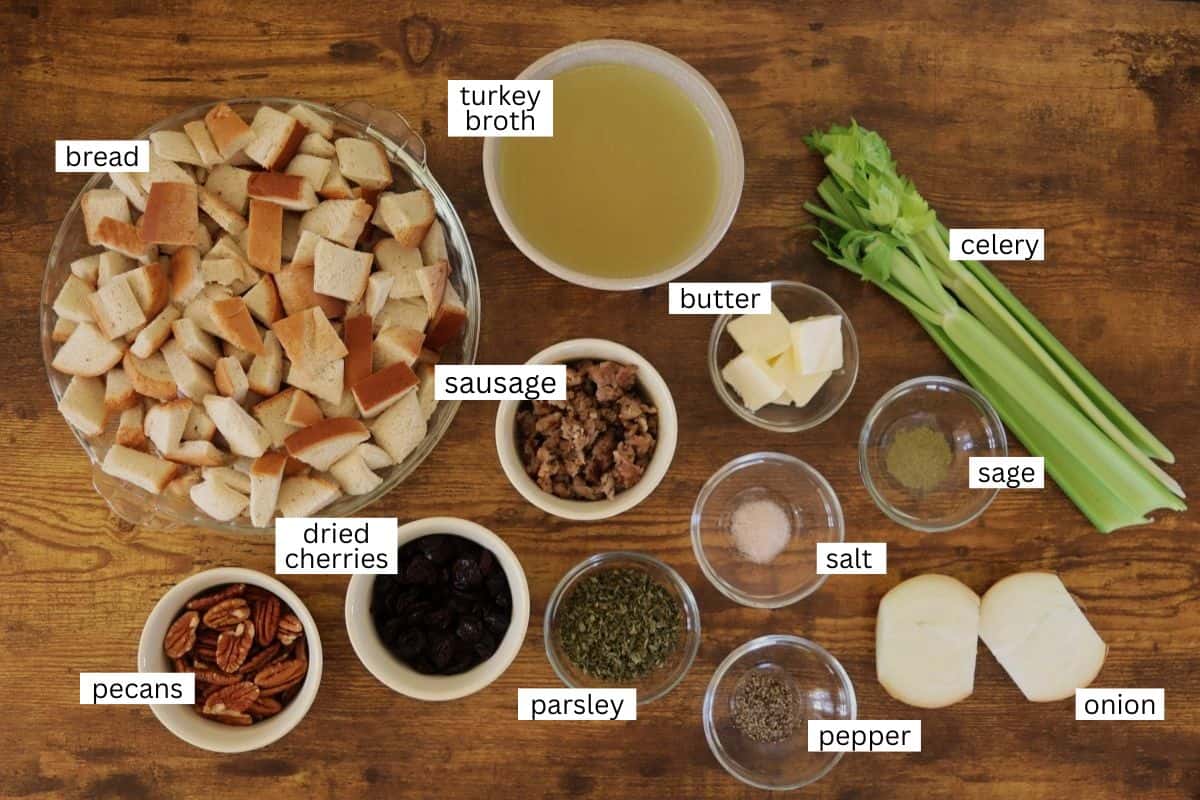 A wooden table with labeled ingredients for stuffing: cubed bread, turkey broth, celery stalks, butter, sausage, dried cherries, pecans, parsley, sage, salt, pepper, and onion.
