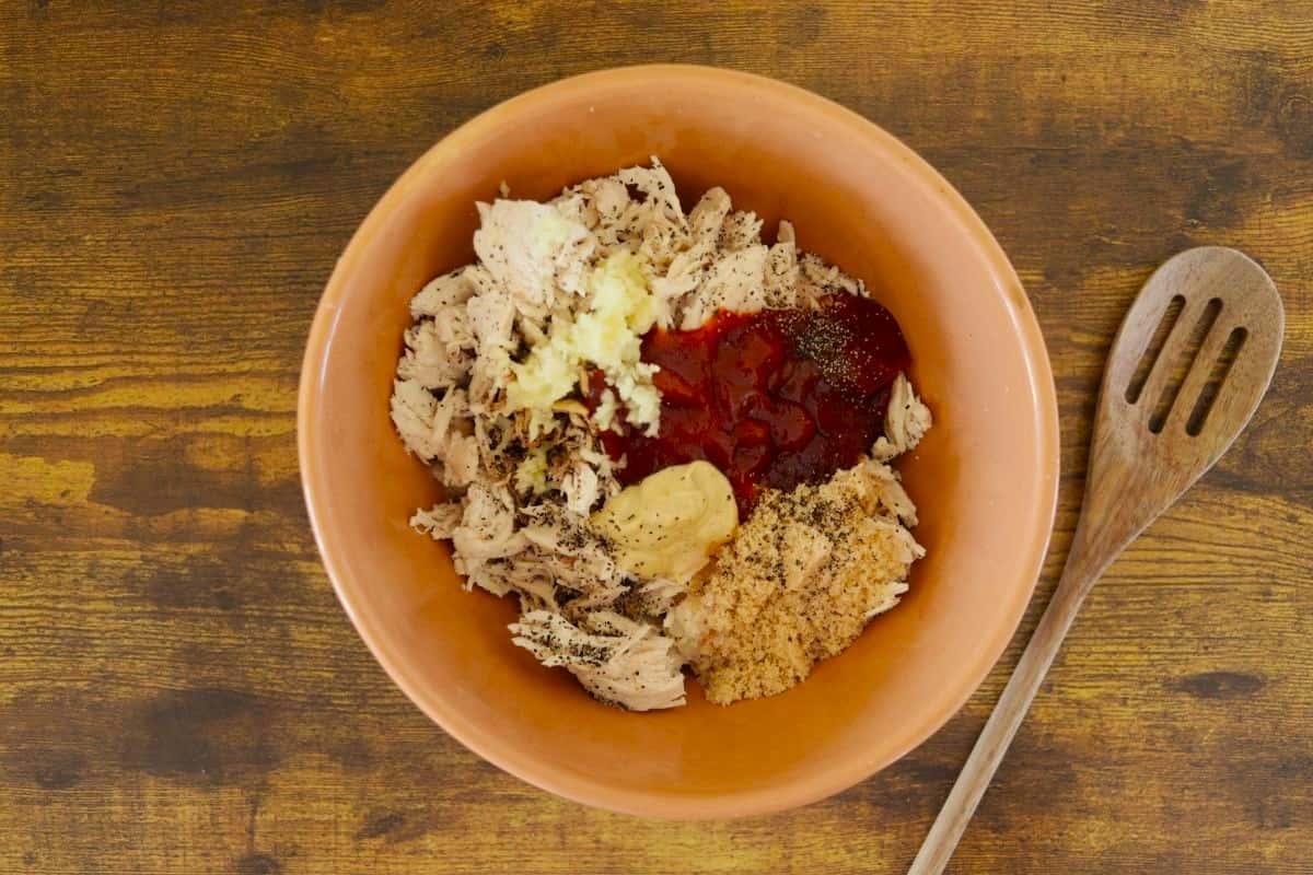 Cooked, shredded turkey in a bowl with ketchup, mustard, brown sugar, garlic, and seasoning visible on top.