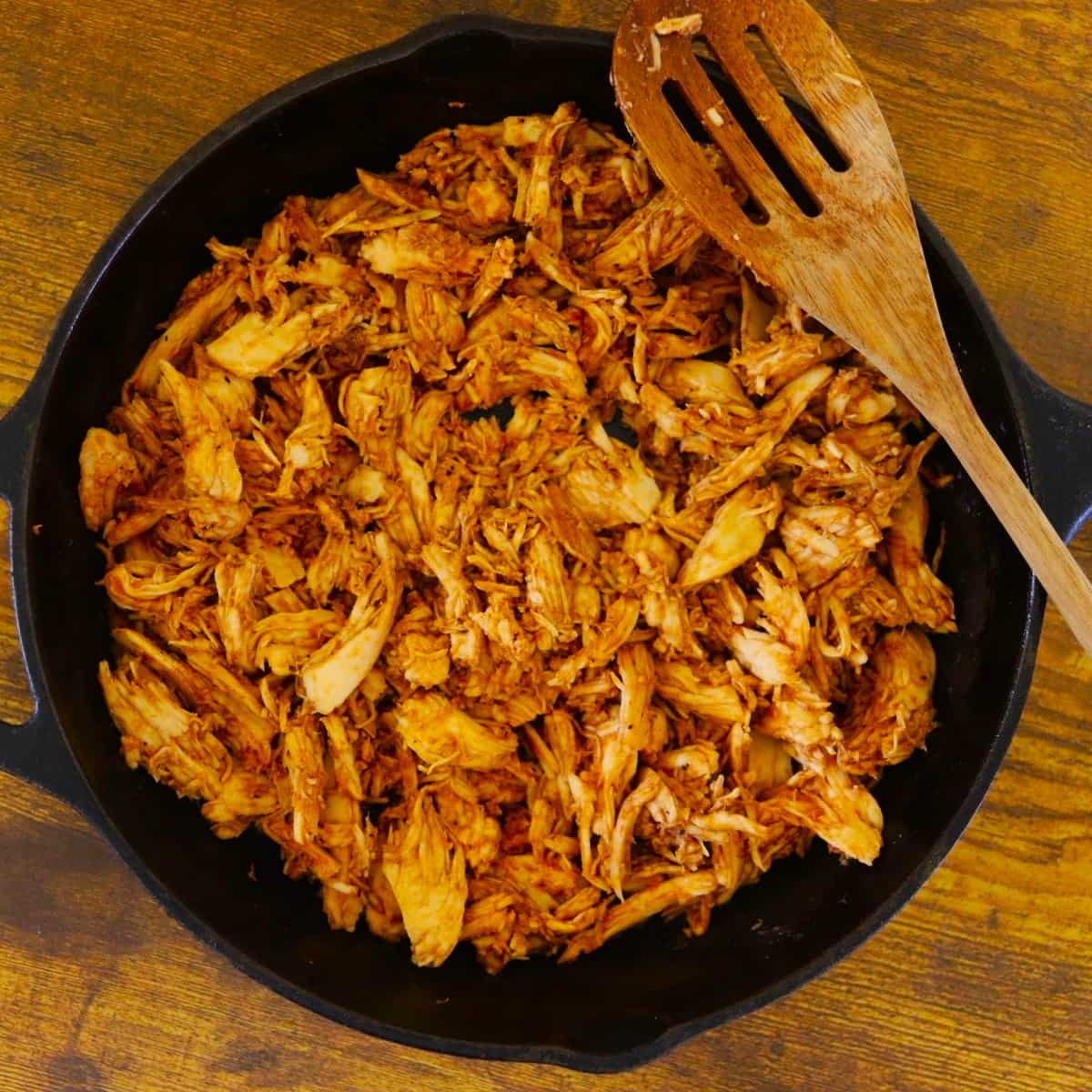 Shredded turkey coated in a reddish brown sauce in a cast iron skillet with a wooden spoon resting on the edge of it.