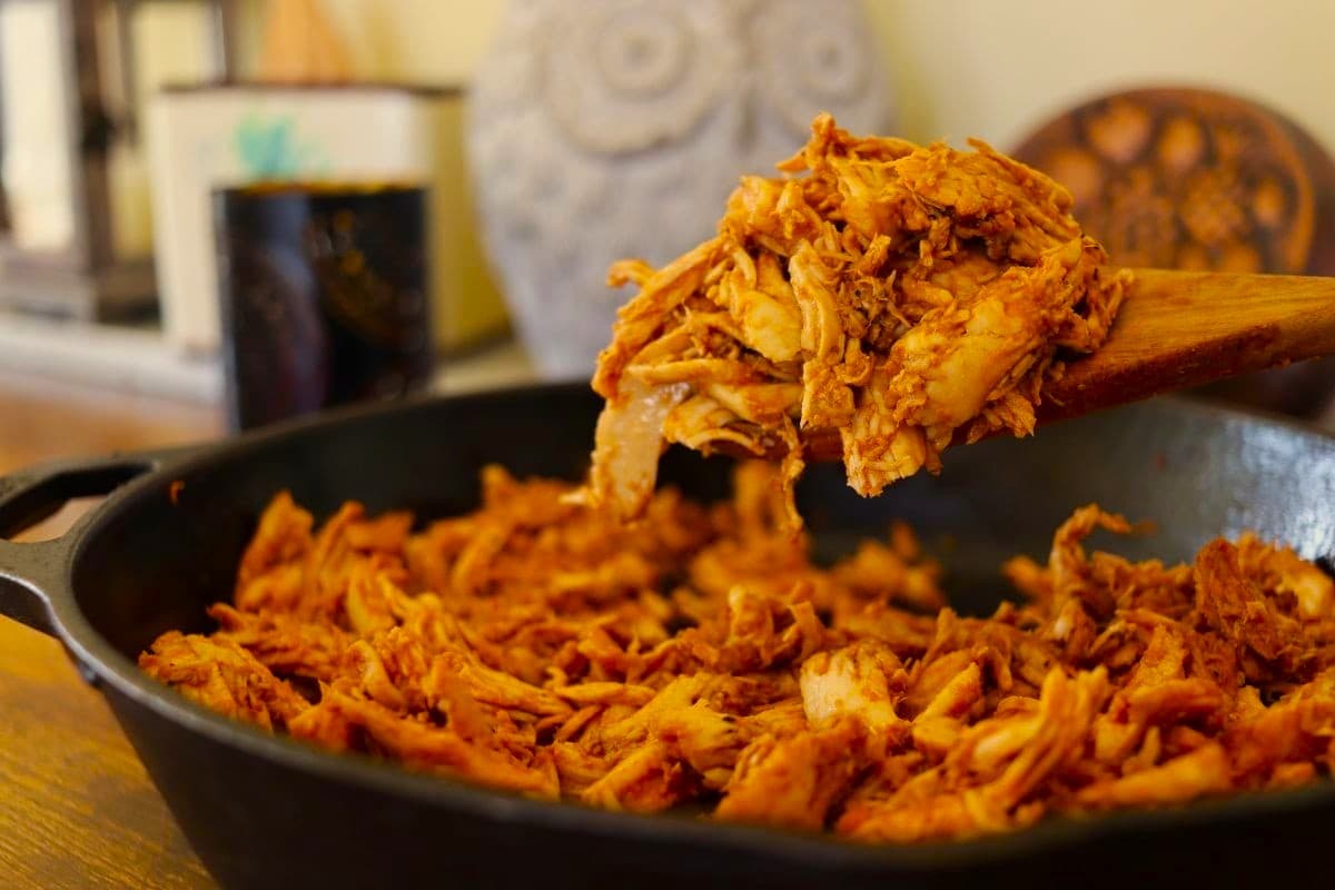 Shredded, seasoned pulled turkey being lifted from a cast iron skillet with a wooden spoon. The chicken is reddish-brown and appears juicy and flavorful. The background is softly blurred with decor visible.
