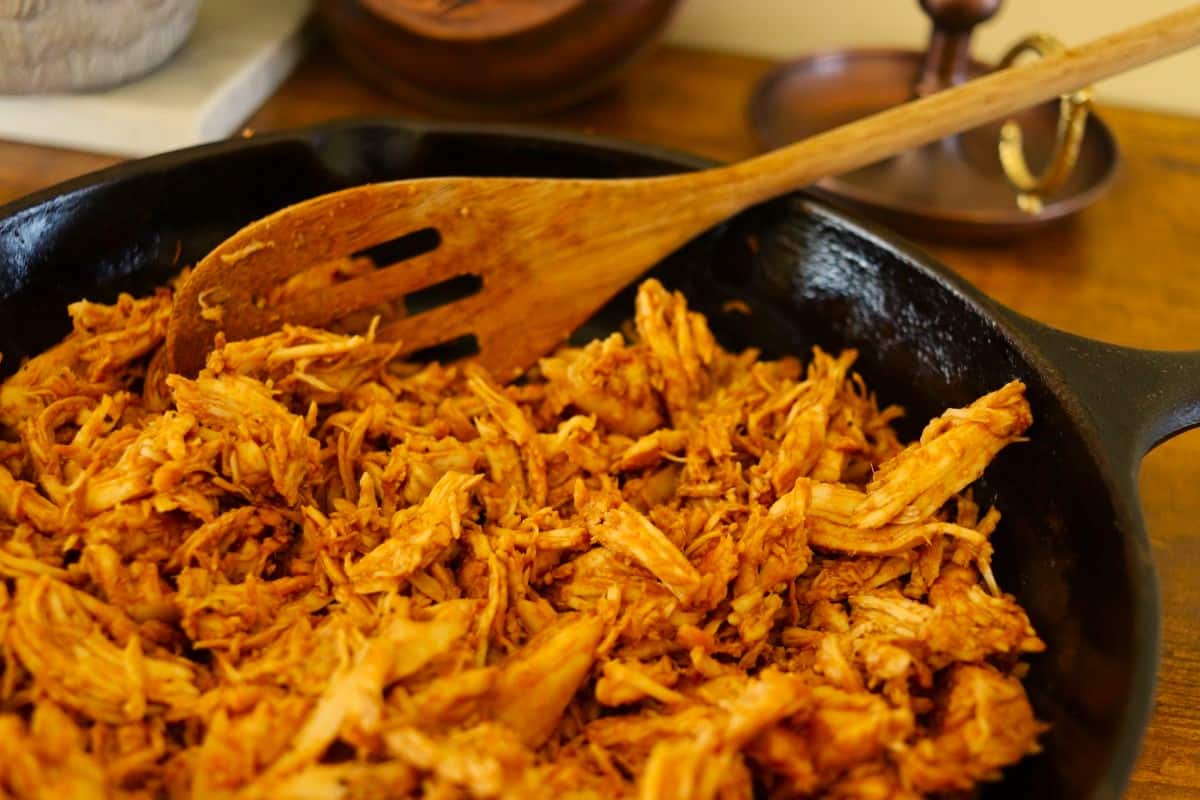 A cast iron skillet filled with shredded, seasoned turkey, with a wooden slotted spoon resting in the pan. The turkey is coated in a reddish sauce.
