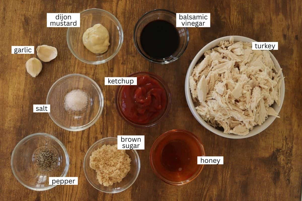 A wooden table displays bowls containing shredded turkey, balsamic vinegar, dijon mustard, ketchup, honey, brown sugar, salt, pepper, and two garlic cloves, each labeled with white text.