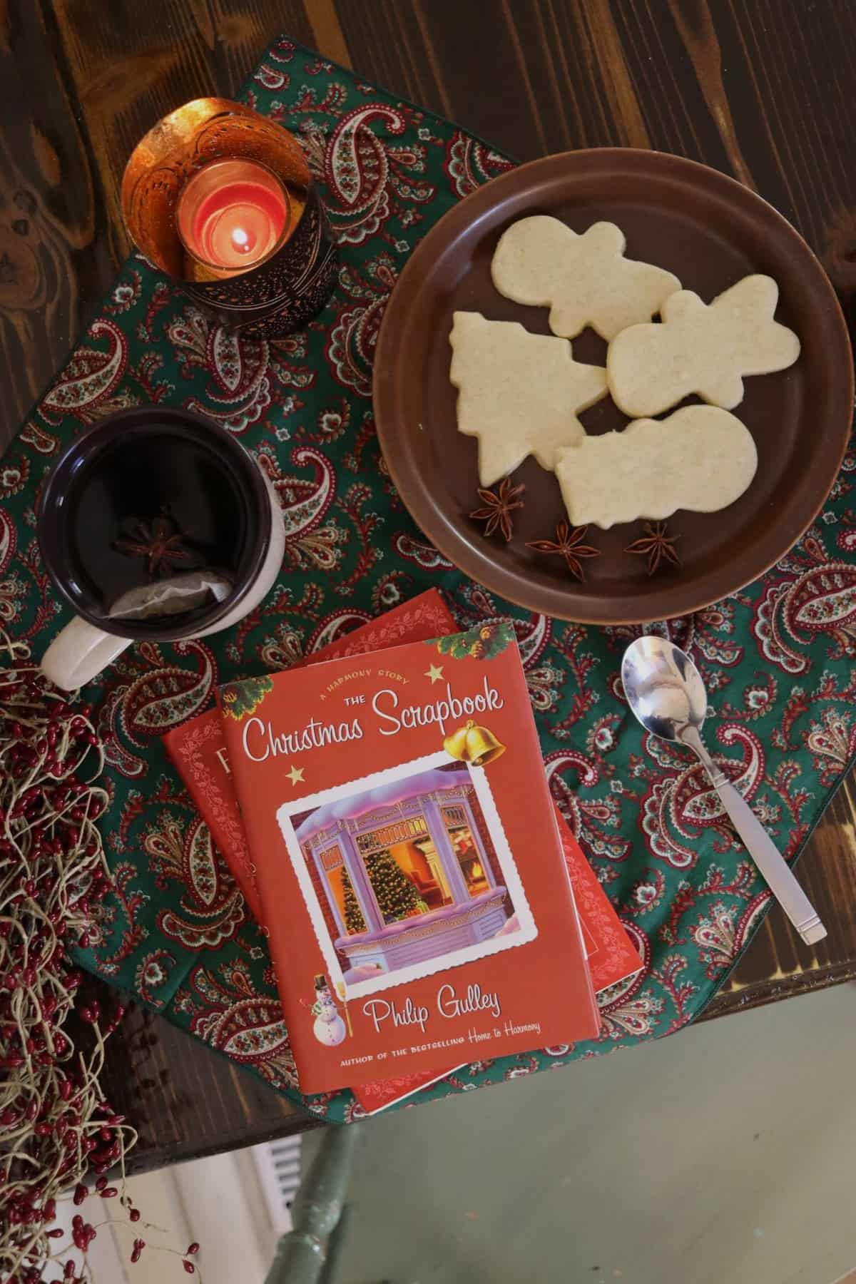 A candle, plate of Christmas shaped cookies, cup of tea, two Christmas books and a spoon on a table with a red and green paisley cloth in the center of it.