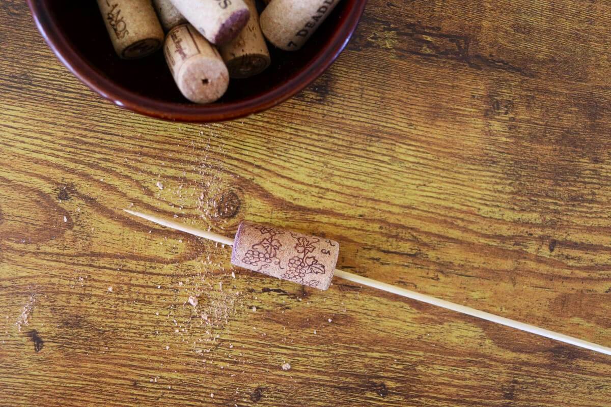 A wine cork on a skewer with cork debris on the table by it and a bowl of wine corks visible in the upper corner of the image.