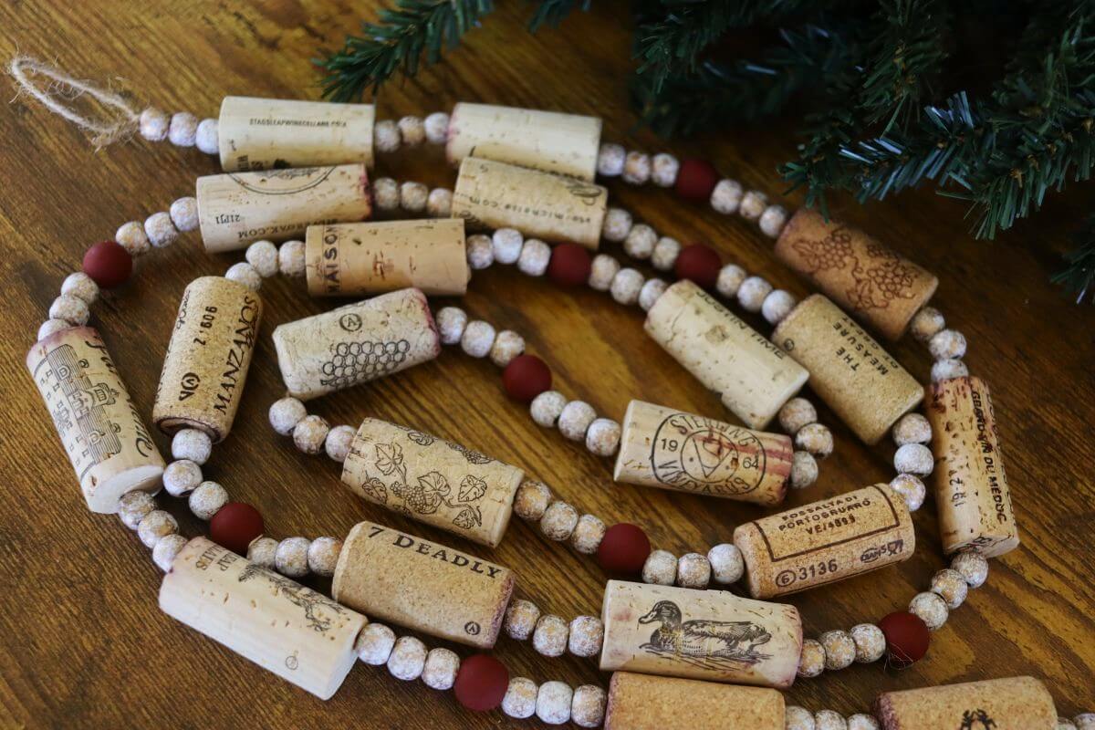 A garland made of wine corks and round beads is arranged on a wooden surface beside green pine branches.