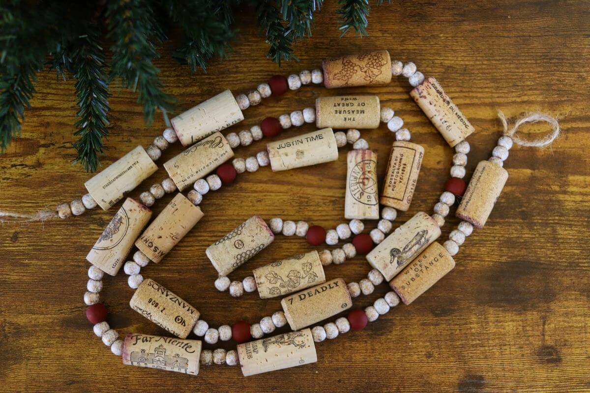 A festive garland made of wine corks, small red and white beads, and twine is arranged on a wooden surface, with green pine branches visible in the upper left corner.