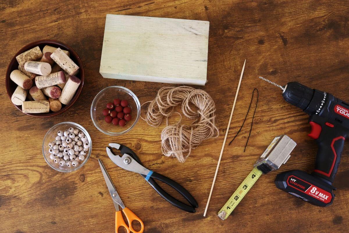 A variety of crafting tools and materials on a wooden surface, including a wood block, corks, string, beads, pliers, scissors, a tape measure, a drill, a skewer.