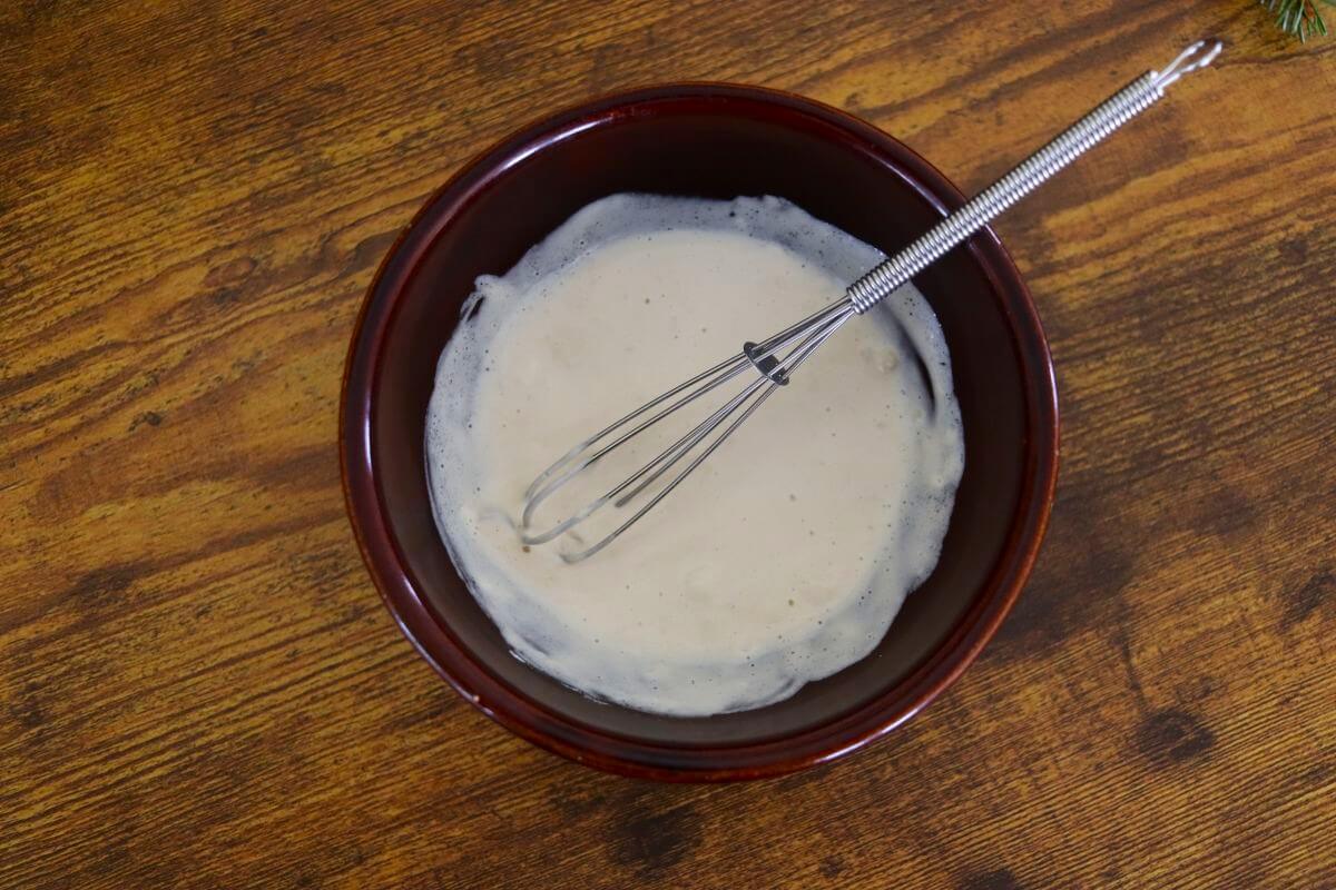 Creamy white cornstarch slurry in a brown bowl with a whisk sitting in it on a wooden surface.