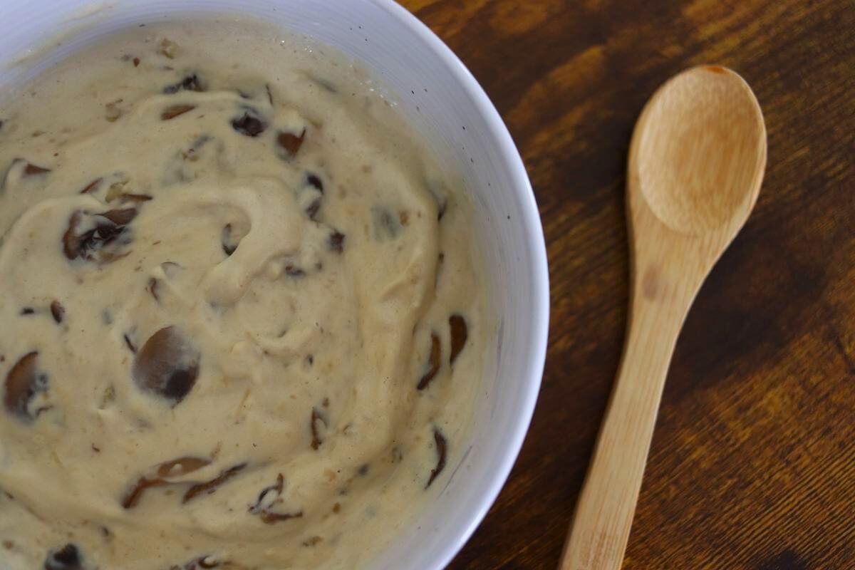 A white bowl filled with creamy mushroom sauce sits on a wooden table next to a wooden spoon. The sauce has visible mushroom pieces mixed into the creamy base.